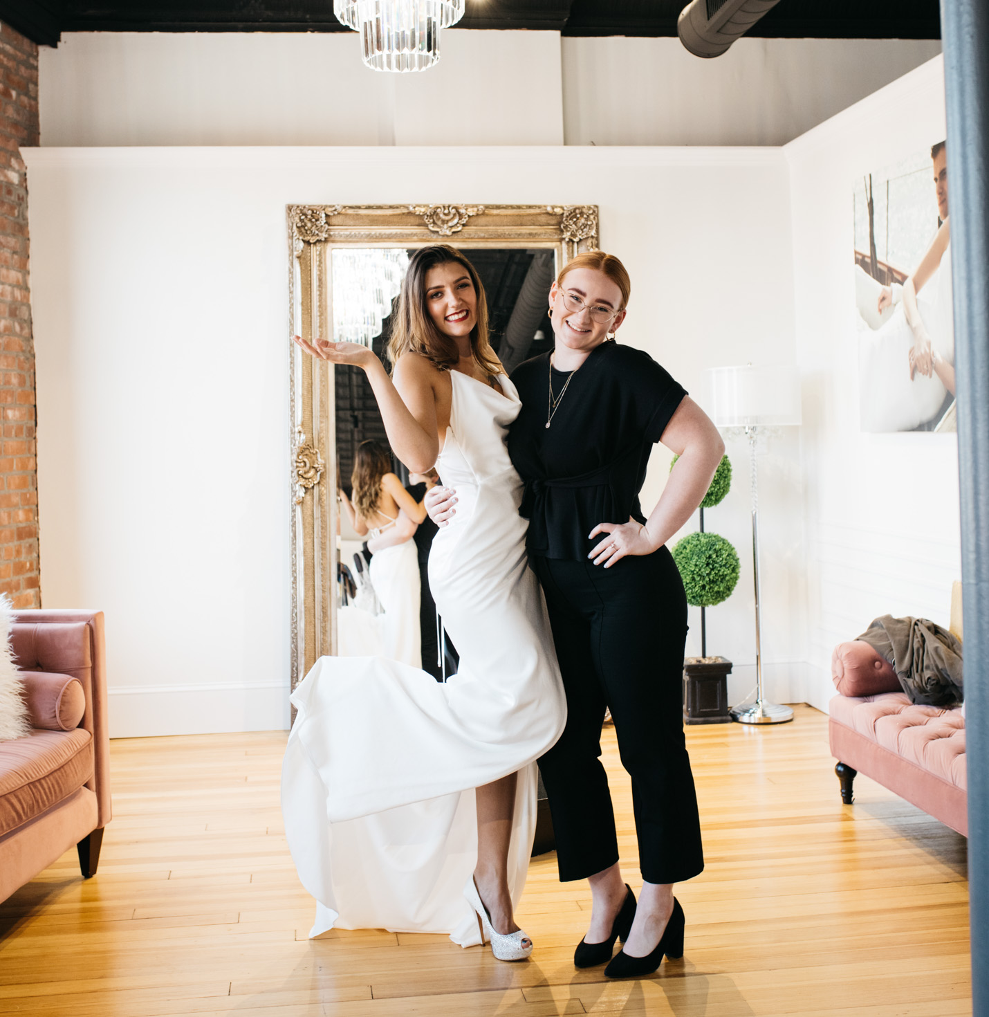 A bride smiling with a store assistant trying on a dress.